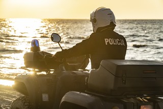 Funkcjonariusz Policji prowadzi czterokołowy pojazd terenowy. Na głowie ma kask. W tle morze i zachodzące słońce.