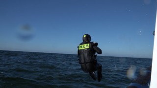 Mężczyzna w pływackim czarnym stroju z pianki z napisem Policja wskakuje do wody. Na głowie ma kask.