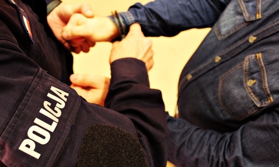 Zdjęcie poglądowe - umundurowany policjant zakłada kajdanki na przeguby dłoni mężczyzny ubranego w niebieską koszulę - sylwetki