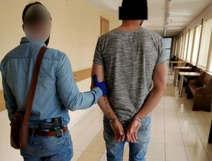 Korytarz. Nieumundurowany policjant  prowadzi mężczyznę z założonymi kajdankami na rękach trzymanych z tyłu. Obaj mężczyźni  są odwróceni tyłem do zdjęcia.