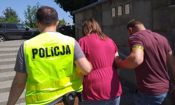 dwaj policjanci prowadzą zatrzymana. Jeden z nich jest w żółtej kamizelce z napisem Policja