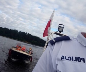 policyjna łódź holuje łódkę z turystami. Z przodu widoczny fragment koszulki policjanta