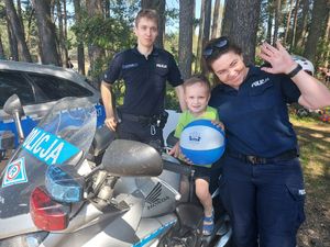 Para policjantów stoi przy motocyklu policyjnym, na którym siedzi mały chłopiec trzymający dmuchaną piłkę.