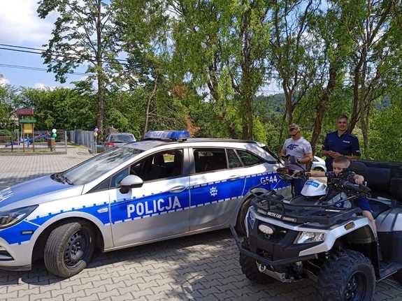 Radiowóz policyjny obok którego stoi kład policyjny, na którym siedzi dziecko obok stoi policjant.