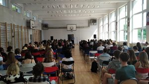 Uczniowie w sali gimnastycznej siedzą na krzesłach ustawionych w rzędach i słuchają prelegenta.