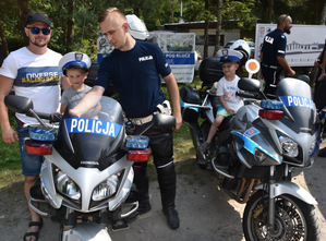 Dwaj chłopcy siedzący na motocyklach policyjnych ubrani w czapki policyjne obok stoi policjant i opiekun dzieci.