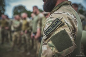 Na pierwszym planie zdjęcie ramienia kontrterrorysty w mundurze ćwiczebnym z naszywką Policja. W nieostrym tle kilkoro kontrterorystów.