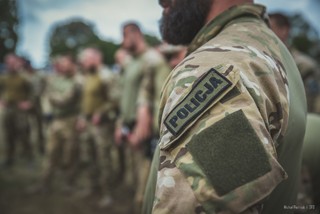 Na pierwszym planie zdjęcie ramienia kontrterrorysty w mundurze ćwiczebnym z naszywką Policja. W nieostrym tle kilkoro kontrterorystów.