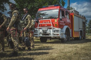 Przed wozem bojowym Straży Pożarnej kilkoro kontrterorystów w pełnym ekwipunku przy ich nogach pojemniki na osobiste liny desantowe.