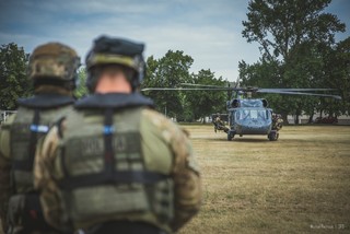 Zdjęcie wykonane zza nieostrych sylwetek dwóch kontrterorystów. W centrum kadru na trawiastym lądowisku stoi policyjny Black Hawk, przy którego burtach kilku kontrterorystów podpiętych linami desantowymi do maszyny.