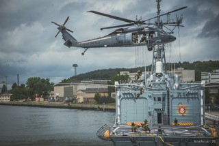 Policyjny Black Hawk w zawisie nad lądowiskiem śmigłowcowym usytuowanym na rufie okrętu Marynarki Wojennej RP T. Kościuszko. Z otwartych drzwi śmigłowca desantują się na pokład okrętu policyjni kontrterroryści. W tle zabudowania portowe.