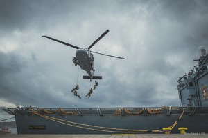 Zdjęcie wykonane z nabrzeża portowego. Policyjny Black Hawk w zawisie nad lądowiskiem śmigłowcowym usytuowanym na rufie okrętu Marynarki Wojennej RP. Z otwartych drzwi śmigłowca desantują się na pokład okrętu policyjni kontrterroryści. W tle zabudowania portowe.