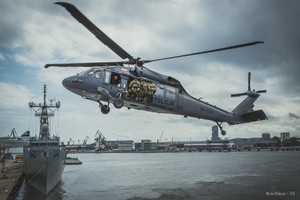Na tle niebieskiego nieba pokrytego chmurami Policyjny Black Hawk z przypiętymi do burty policyjnymi kontrterrorystami podczas podchodzenia do miejsca desantu. W tle dziób okrętu Marynarki Wojennej RP, nabrzeża oraz żurawie portowe.
