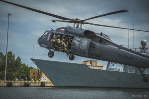 Policyjny Black Hawk lecący nad akwenem na tle lewej burty okrętu Marynarki Wojennej RP. W otwartych drzwiach bocznych śmigłowca podpięci linami desantowymi policyjni kontrterroryści.