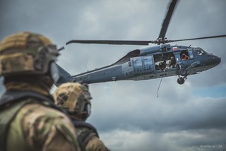 Na pierwszym planie dwie sylwetki policyjnych kontrterorystów w centrum kadru przelatuje policyjny Black Hawk z otwartymi drzwiami bocznym, z których wychylają się policyjni instruktorzy oraz crew chief.