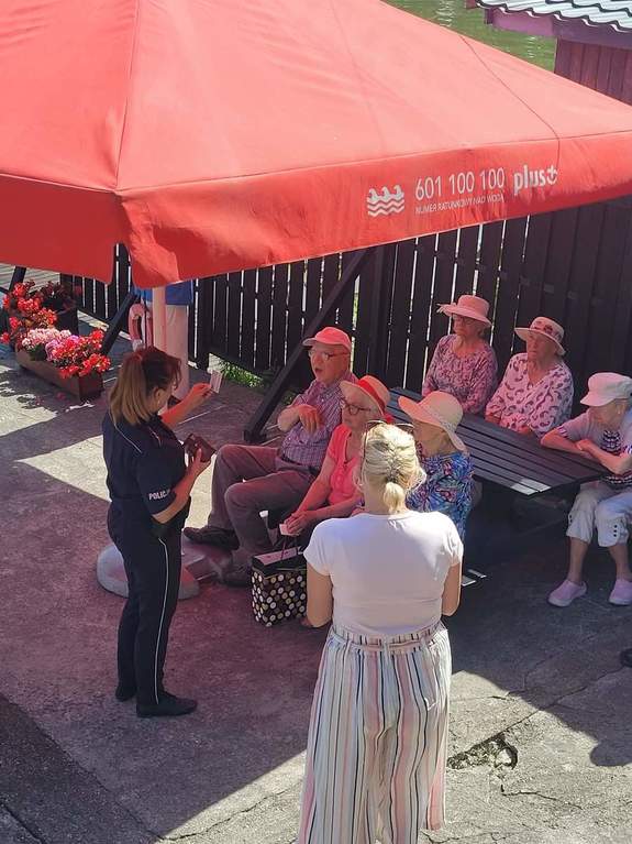 Policjantka rozmawia z seniorami siedzącymi pod namiotem obok stoi ich opiekunka.