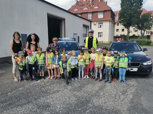 Policjant pozuje do zdjęcia z grupą dzieci i ich opiekunkami za dziećmi stoją dwa radiowozy. Dzieci mają na sobie kamizelki odblaskowe.