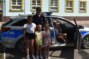 Policjantka stoi z trzema dziewczynkami przy radiowozie policyjnym, jedna siedzi w środku. Wszyscy pozują do zdjęcia.
