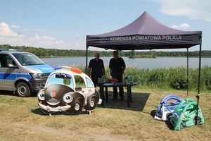 Dwóch policjantów stoi pod namiotem profilaktycznym. Po lewej stronie stoi radiowóz w tle zbiornik wodny.