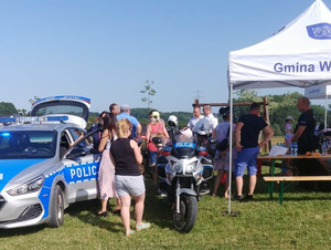 Policjantka stoi przy radiowozie po jej prawej stronie stoi motocykl policyjny i namiot, wokół przemieszczają się uczestnicy imprezy.