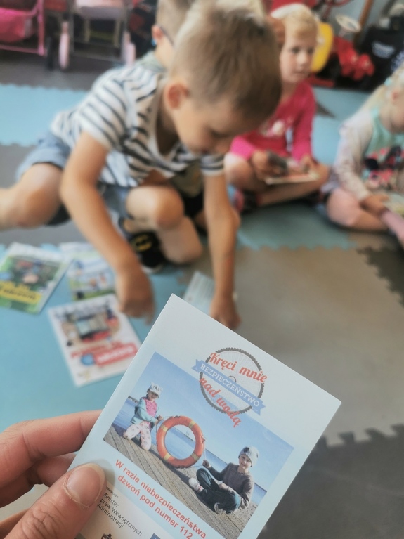 Na pierwszym planie ulotka akcji Kręci mnie bezpieczeństwo nad wodą w tle grupa dzieci siedzących na podłodze.