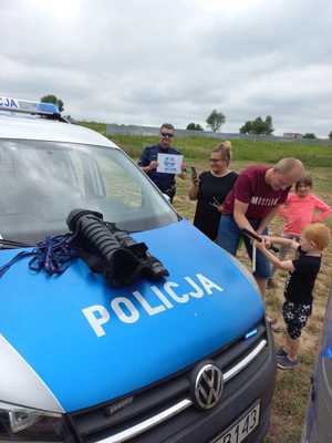 Radiowóz policyjny po jego prawej stronie uczestnicy festynu i policjant trzymający plakat akcji Kręci mnie bezpieczeństwo nad wodą.
