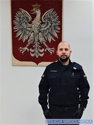 policjant w mundurze a za nim godło na ścianie