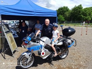 Dziewczynka na motocyklu policyjnym obok stoi policjant, w tle namiot profilaktyczny i drzewa.