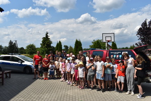 Grupa dzieci pozuje do zdjęcia z opiekunami, policjantami i ratownikami medycznymi. W tle radiowóz i pojazd ratowników medycznych.