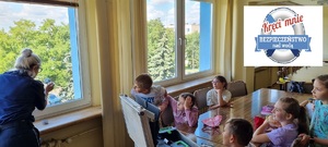 Policjantka pokazuje grupie dzieci jak ujawnić odciski palców na szybie okna, w prawym górnym rogu logo akcji &quot;Kręci mnie bezpieczeństwo nad wodą&quot;.