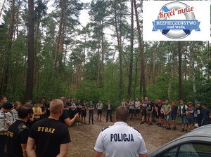 Na polanie leśnej grupa harcerzy ustawionych w okręgu rozmawia ze strażakami i policjantem. W tle wysokie drzewa,  w prawym górnym rogu logo akcji &quot;Kręci mnie bezpieczeństwo nad wodą&quot;.