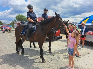 Dwa konie służbowe policji, na których siedzą policjanci. Przed nimi stoi grupa dzieci głaskających konie. W tle wozy strażackie.