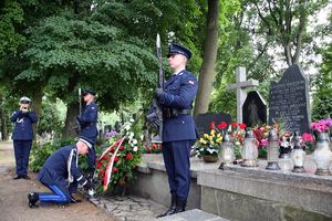 Komendant Główny Policji kuca przy wieńcu złożonym przy grobie i ustawia znicz. Z boku, przy grobie dwaj policjanci pełnią wartę honorową, Widać również grającego na trąbce funkcjonariusza