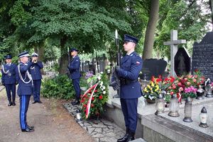 Komendant Główny Policji stoi przed grobem i salutuje. Z boku, przy grobie dwaj policjanci pełnią wartę honorową, z boku widać grającego na trąbce policjanta