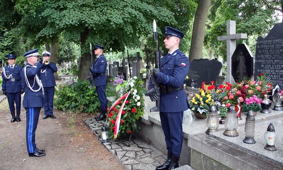 Komendant Główny Policji stoi przed grobem i salutuje. Z boku, przy grobie dwaj policjanci pełnią wartę honorową, z boku widać grającego na trąbce policjanta