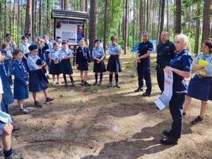 Policjantka rozmawia z grupą harcerzy ustawionych w okręgu, w tle wysokie drzewa.