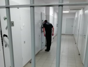 policjant dozorujący zatrzymanego przez wizjer w drzwiach celi. Na pierwszym planie kraty