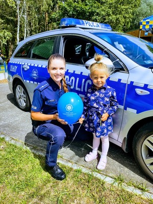 Policjantka trzyma balonik obok stoi dziewczynka, w tle radiowóz policyjny.