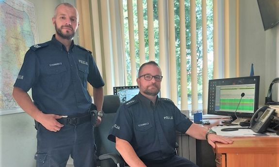 dwaj umundurowani policjanci - jeden stoi, drugi siedzi na krześle