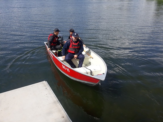 Łódka strażacka płynie w kierunku brzegu, w łódce siedzi policjant i dwóch strażaków.