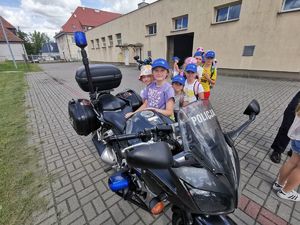 Dziewczynka stoi obok motocykla policyjnego, za nią dzieci ustawione w rzędzie czekają na swoją kolej.