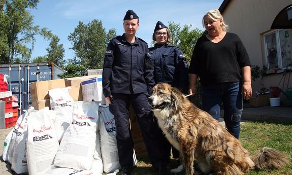 dwie policjantki stoją z kobietą ubraną po cywilnemu, przed nimi siedzi pies, a z boku widać worki