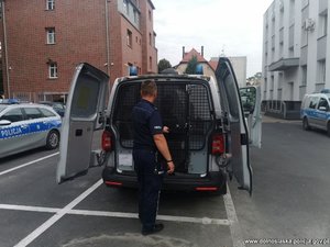 policjant stoi przy radiowozie policyjnym, w którym znajduje się zatrzymana osoba