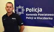 policjant stoi na tle ściany, na ścianie wisi banner z napisem Policja Komenda Powiatowa Policji w Kluczborku