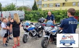 Dwa motocykle policyjne obok nich stoją policjanci i grupa dzieci z opiekunem.