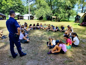 Policjant rozmawia z grupą dzieci siedzących na polanie.