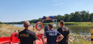 Policjant i dwóch strażaków na brzegu akwenu wodnego.