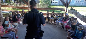 Policjant rozmawia z dziećmi siedzącymi przed nim na ustawionych w okręgu krzesłach.