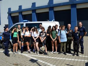 Harcerze pozują do zdjęcia z policjantami przed budynkiem Komendy Miejskiej Policji w Płocku za nimi stoi radiowóz.
