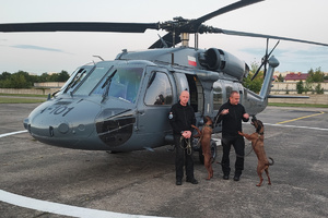 Dwaj policjanci z psami służbowymi, które stoją na dwóch łapach obok policyjnego śmigłowca na lądowisku.